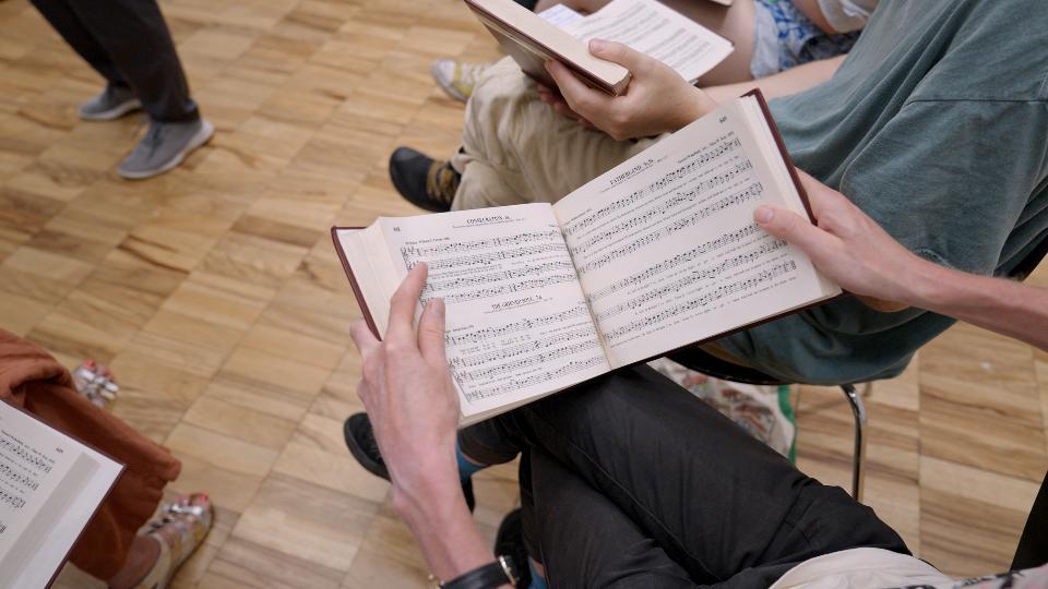 Looking over the shoulder of a seated singer we see the Sacred Harp book open, propped on their knee and held in both hands. It's a large book, with rows of shapenote music and song lyrics. printed on the pages.