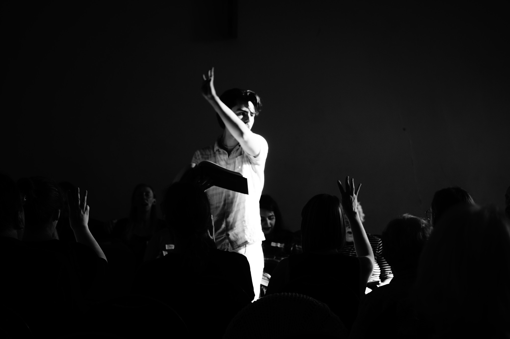 A leader stands in the centre of the square of seated singers, with his book open. He is signalling '3rd verse' with his hand swept up in the air, and others are echoing the gesture. The image is in deep shade but he is brightly lit from the right, hinting at the low autumn sun streaming from the large window. It is a very dramatic black and white shot.