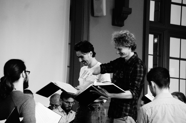 Two leaders stand side by side, facing seated singers. They're looking at their open tunebooks and laughing together. 