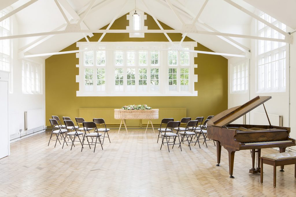 A large bright room with high ceiling, wooden floor and large window. The focal point is a willow coffin decorated with flowers. Rows of chairs face towards it as if set up for a funeral service. The image gives a feeling of serenity and peace.