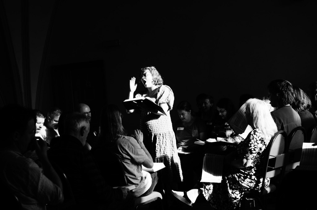 A leader stands, holding a large book open on her arm. Her right hand is raised, beating time and she is singing with passion. There are seated singers around her, all in shadow. She is lit by bright sun from the right, making the photo sharply contrasted in black and white.
