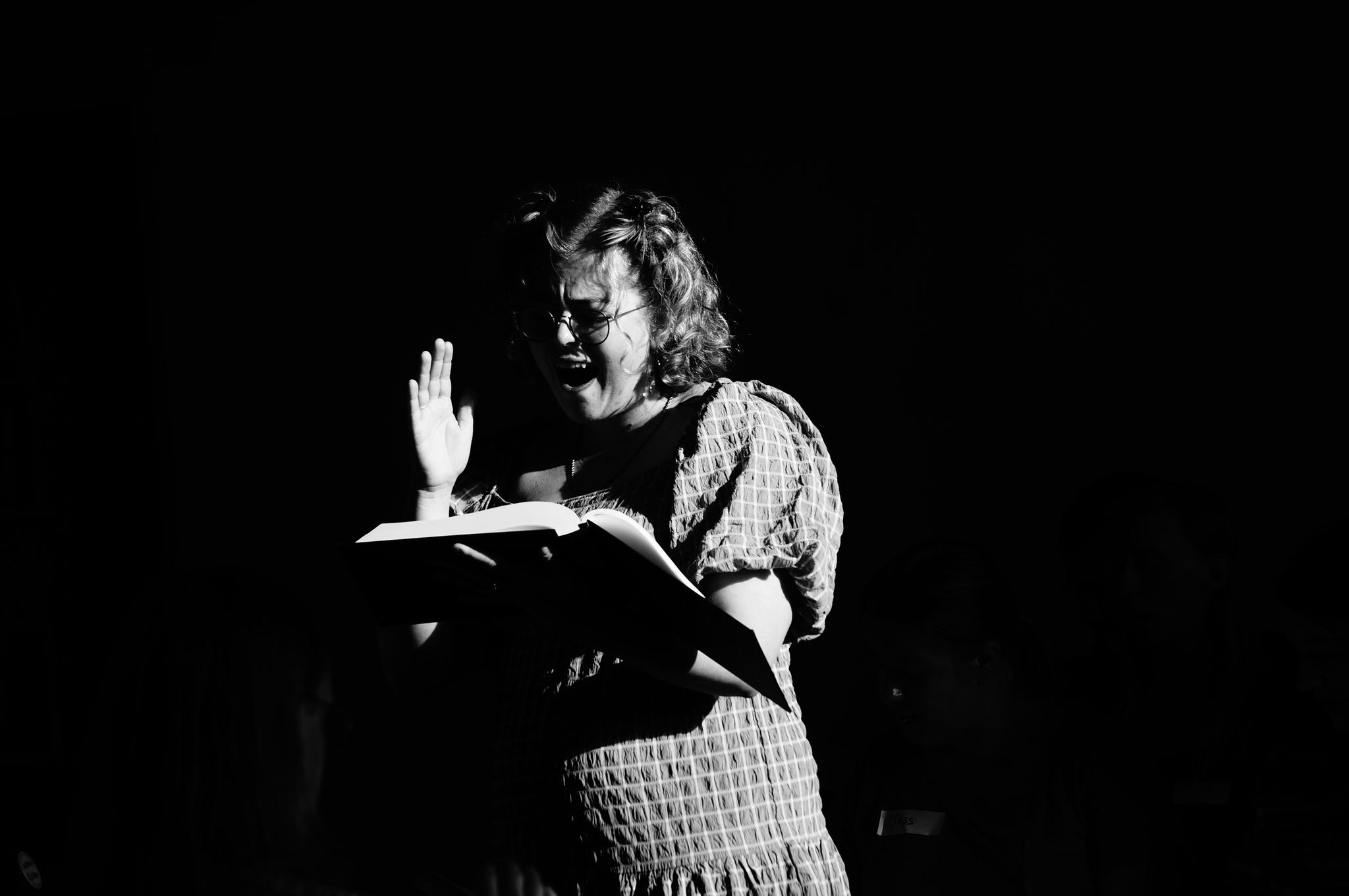 Sophie Lockhart, who will chair the 2025 All-Day. The image is an atmospheric black and white image showing a young white woman with shoulder-length curly hair holding her Sacred Harp book and singing with passion. Her right hand is held in the air, as she beats the time of the song she's leading. 