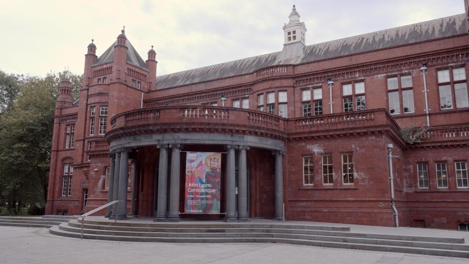 The impressive front entrance to The Whitworth art gallery. A red brick and terracotta building. It is two-storeys high with rows of windows and a raised semi-circular entrance with grey pillars which looks very grand.