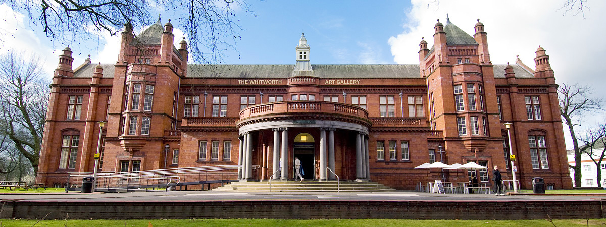 The impressive front entrance to The Whitworth art gallery. A red brick and terracotta building, lit up by the sun so it's firey orange. It is two-storeys high with rows of windows and a raised semi-circular entrance with grey pillars which looks very grand.