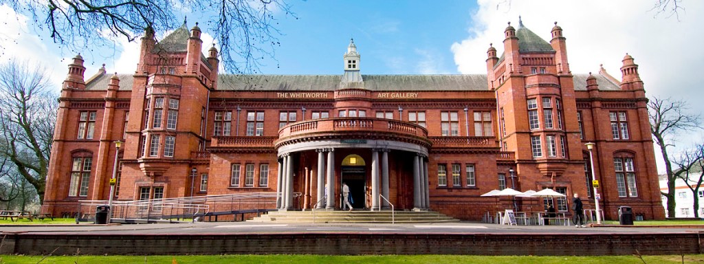 The impressive front entrance to The Whitworth art gallery. A red brick and terracotta building, lit up by the sun so it's firey orange. It is two-storeys high with rows of windows and a raised semi-circular entrance with grey pillars which looks very grand.