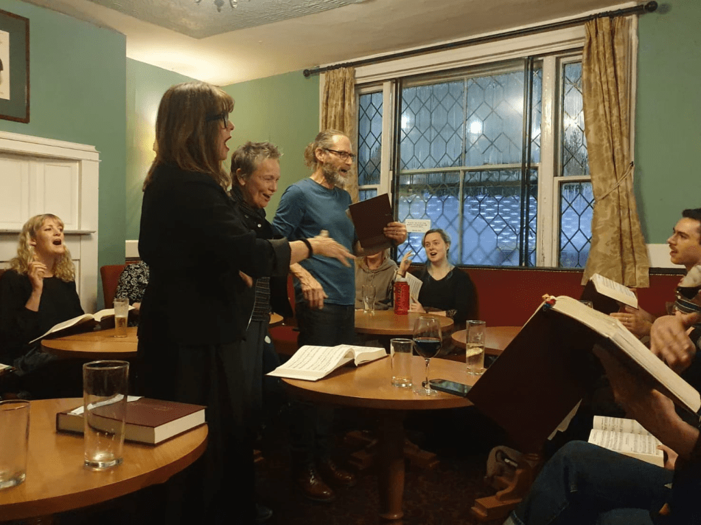 Three singers stand, singing from the Sacred Harp. They are surrounded by seated singers, all looking attentively and singing with passion. They're hemmed in by pub furniture. It looks cosy - a low glow of amber light and the lattice windows are steamed up. 