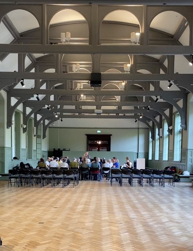 A group of singers sitting in a square with a leader in the centre, his hand raised and a book resting on his other arm. The image is taken from a distance, so the foreground is a long expanse of wooden floor. The image is taken in the Grand Hall at The Whitworth. It is beautiful - soft light, soft green walls and darker green arched wooden supports fitted into the high ceiling. 
