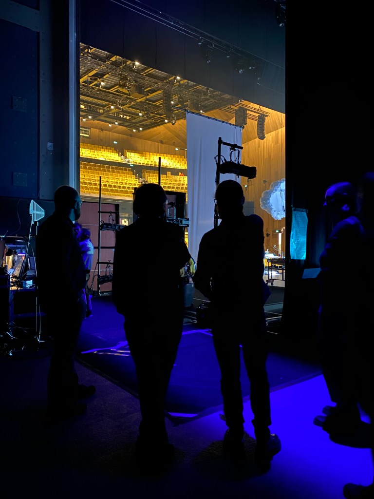 Four figures in shadow wait backstage, in a blue-tinged near-darkness. Beyond them we can see the theatre auditorium with hundreds of golden yellow velvet seats. There is rigging and props on the stage. 