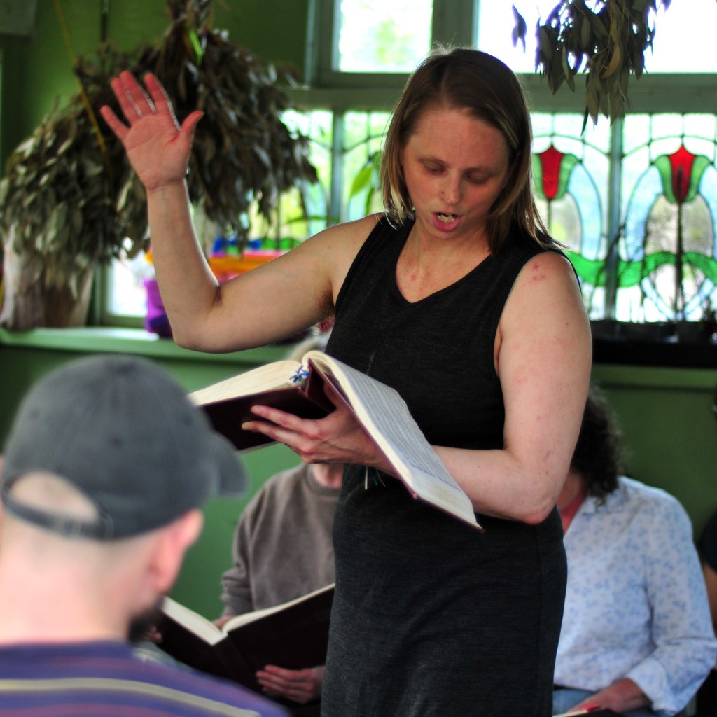 Hannah Land leading a song at the Frivolous Fields festival at Platt Fields in June 2025. Mika Upton (baseball cap) visible in the foreground