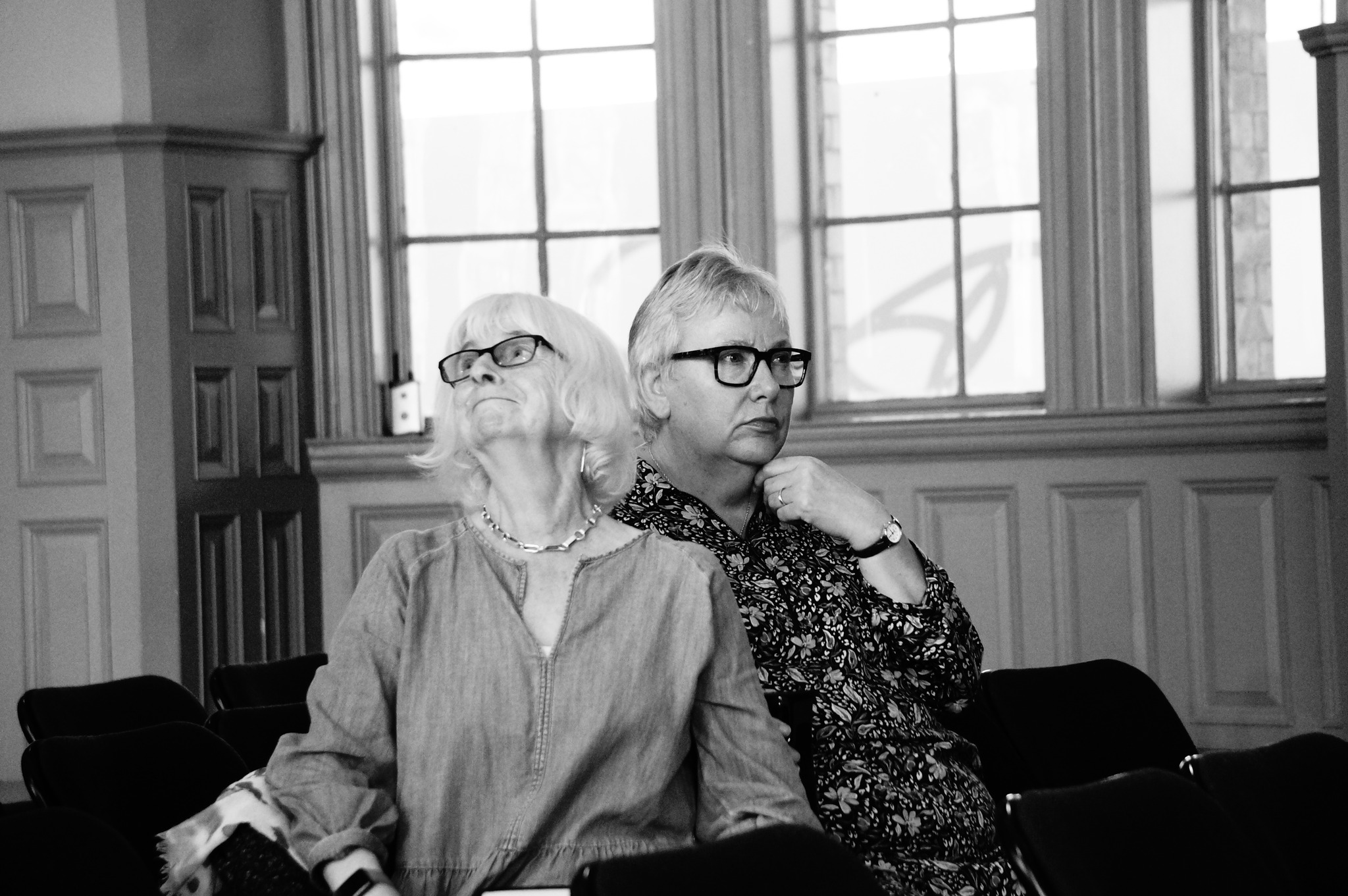 A black and white image of two singers sitting together but twisting away from each other.. The two women's heads are close, yet looking in opposite directions. One looks to the ceiling with a faint smile. The other, with her hand to her chin, is frowning and looks pensive, almost stern.