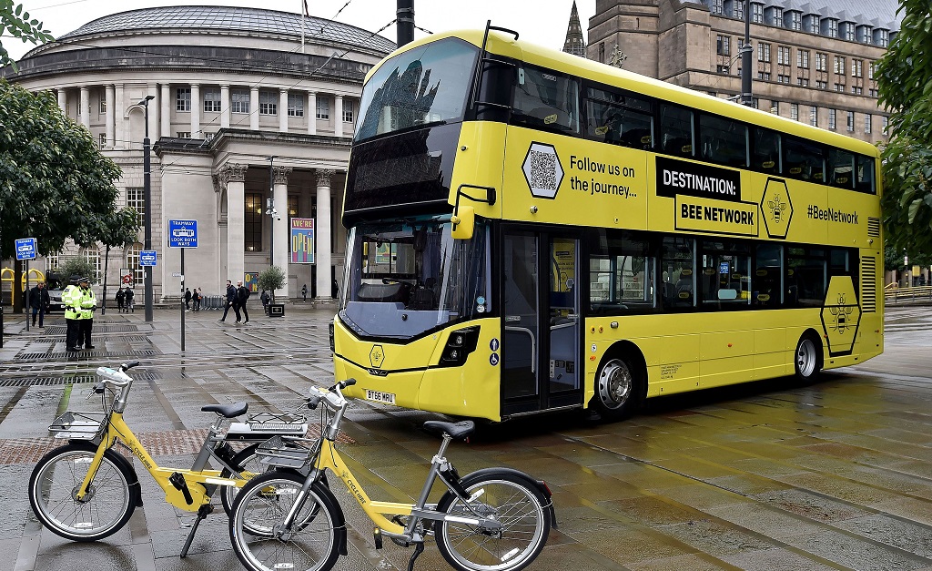 A promotional image from Transport for Greater Manchester showing elements of the Bee Network. A new yellow electric bus and two yellow hire bikes in the foreground. Behind is the circular Central Library. 