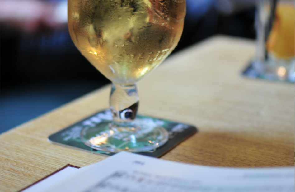 We can see the lower part of a glass of lager. The European pint glass has a stem and is covered in condensation, suggesting a refreshing cold drink on a warm day. In the foreground we see the corner of a Sacred Harp book which is open on the table in front of the drink.