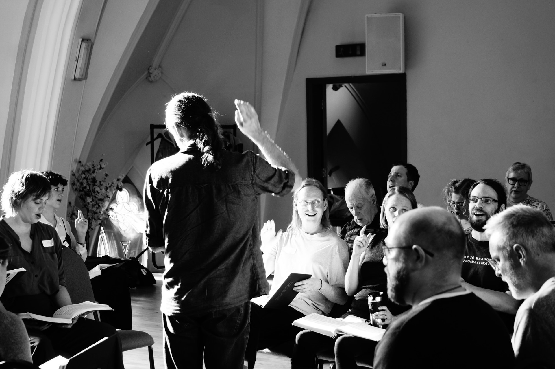 A black and white image from an All-Day singing. A man stands in the foreground of the image, book in hand and arm raised to beat the time of the song he is leading. Beyond him sits several rows of singers, books open and singing. There's strong autumnal sunlight slanting through the window to the left, lighting up their faces. 
