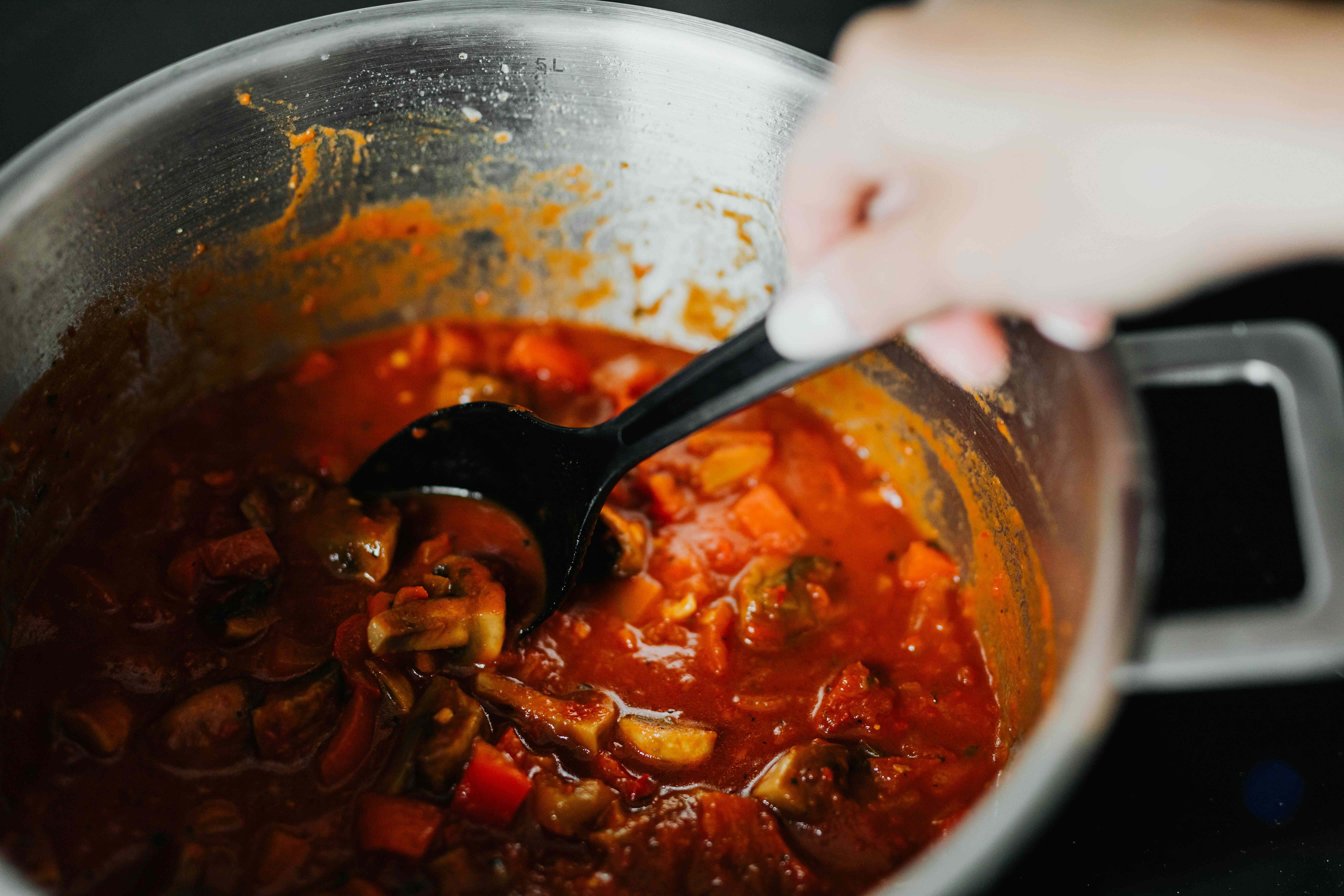 A generic image of a very large metal pan and a hand holding a spoon which is stirring the contents - a delicious-looking rich tomato and vegetable stew. 