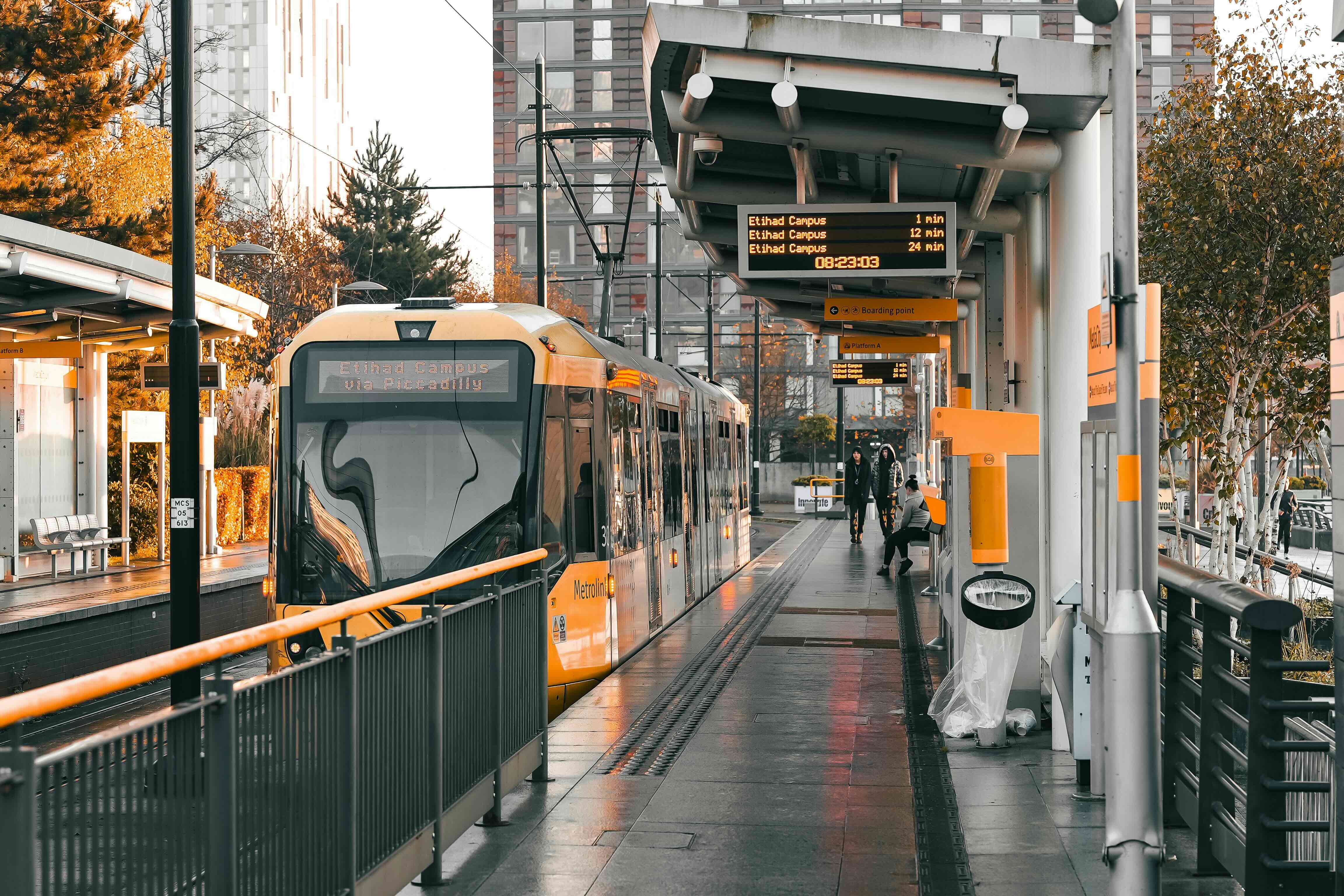 An image of a Metrolink tram taken from the platform. It shows the front of the yellow tram and the display board says Etihad Campus.