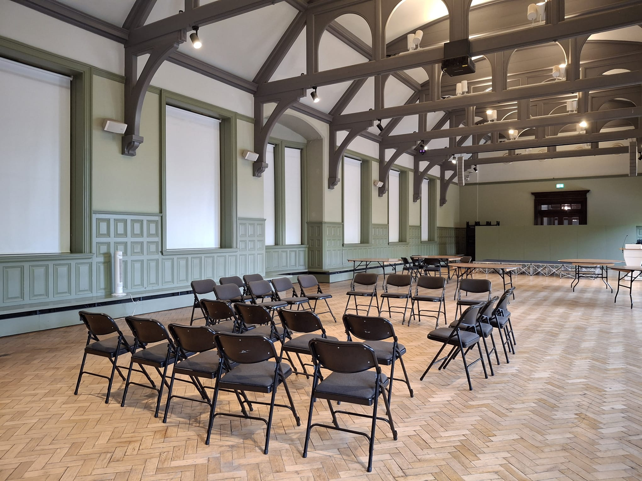 The Grand Hall at The Whitworth. Black fold-out chairs are arranged in a hollow square in the centre of the room. There are no people in the image. There is a wooden floor in a beautiful herringbone pattern, soft green walls and darker green arched wooden supports fitted into the high ceiling. 