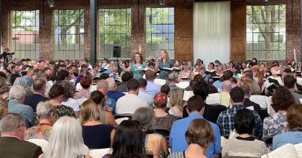 Vicki Elliott and Samantha Cole, both in greeny blue dresses, their arms raised, standing in the middle of a huge crowd of seated singers in a hall in Atlanta