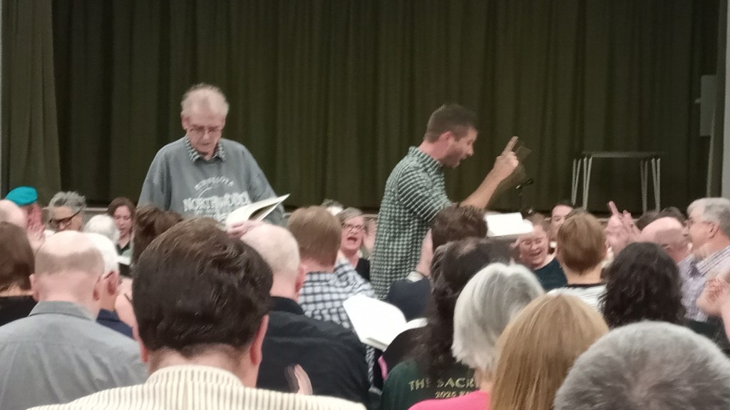 Chris Brown and Matthew Parkinson leading Gwehelog. They are standing in the middle of the square, surrounded by singers, with a dark green curtain in the background. Matthew is seen sideways on, facing the trebles, and raising one finger; some of the trebles mirror his gesture