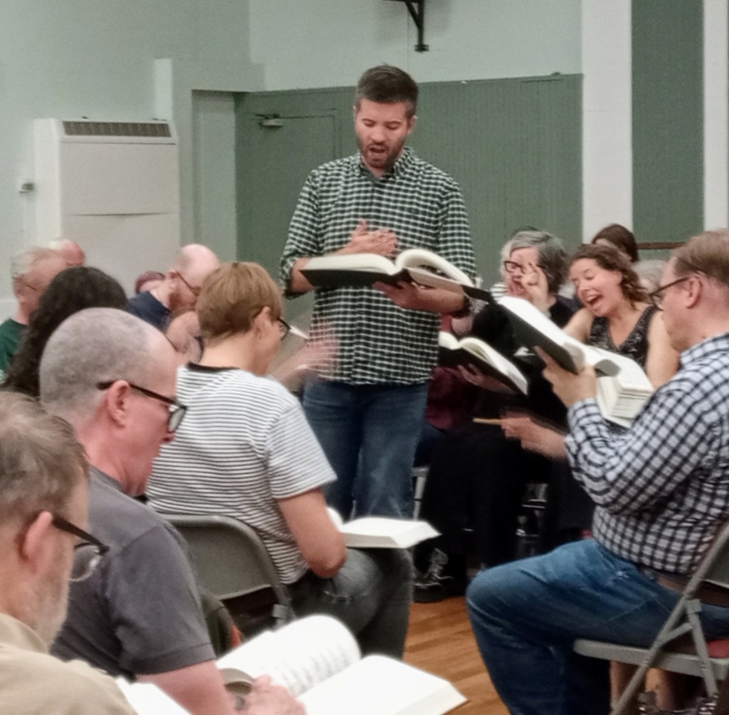 Matthew Parkinson leading He Hath Saved Us on the Friday evening of the UK convention, 2025. He is wearing a green checked shirt and jeans, and his right hand seems to be gesturing at his heart as he holds the book in the left. He is surrounded by singers, with the altos visible behind him