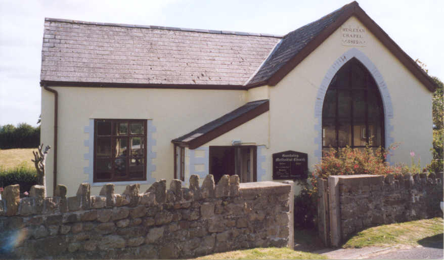 A small chapel in Gwehelog, with creamy walls and a slate roof. There is a sign reading "Wesleyan Chapel 1822" over a large arched window, and a wooden board reading "Gwehelog Methodist Church" by the door