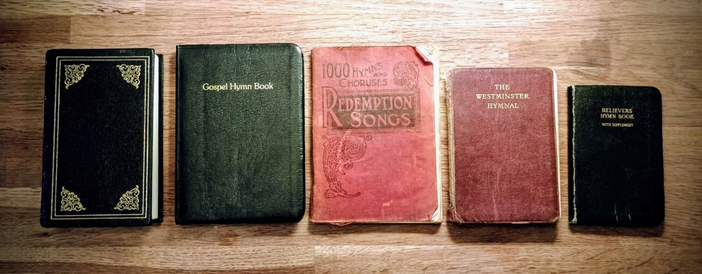 A row of five hymn books - a black one with gold borders, title invisible, but it must be Primitive Hymns; a black one, labelled "Gospel Hymn Book" in gold; a very battered red one, titled "Redemption Songs: 1000 Hymns and Choruses"; another red one, "The Westminster Hymnal"; and a small black one, "Believers Hymn Book with supplement".