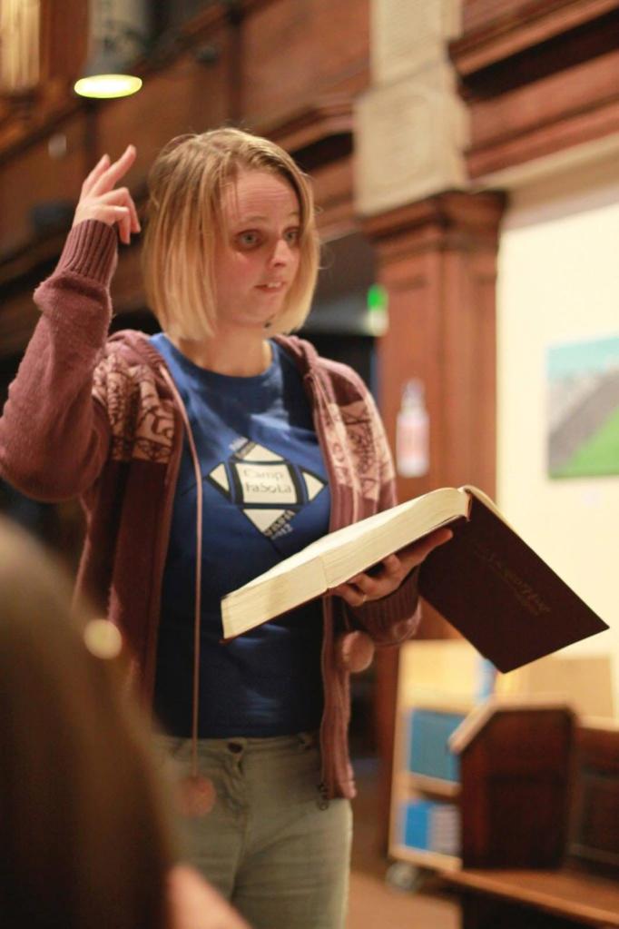 Hannah leading at Sacred Trinity Church, in close-up. She is wearing a Camp Fasola t-shirt; her right arm is raised, her left is holding the book.