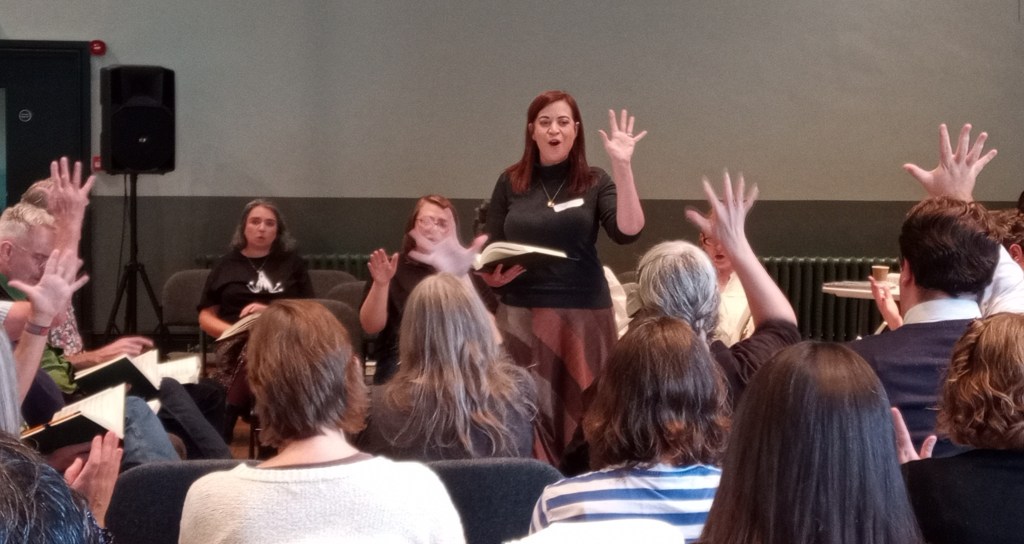 Susanna standing on the middle of the square; she is holding up her right hand with all five fingers splayed to indicate that we should sing the fifth verse, and several singers are mimicking her by raising their own open hands
