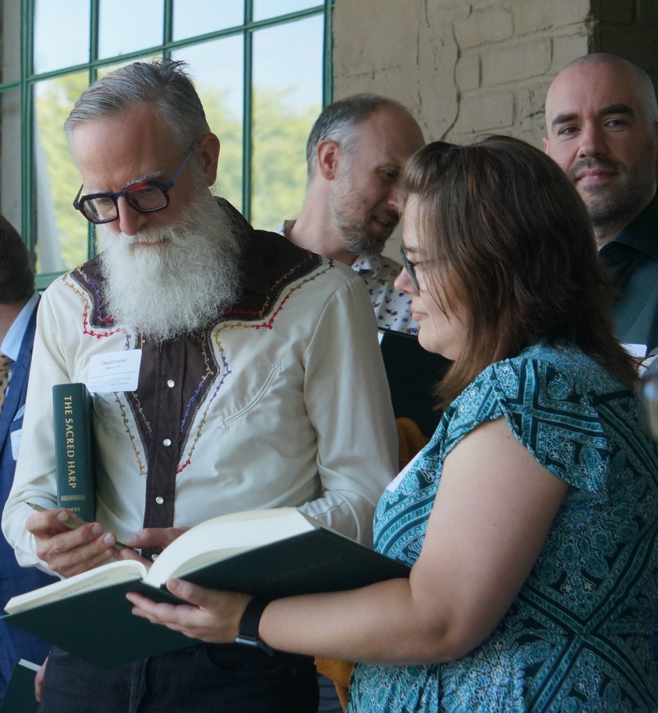 David Smead, a man with a long white beard and a copy of The Sacred Harp 2025 under his arm, signing the page with his own song in Vicki's copy of the book. Vicki is wearing a turquoise and blue dress. Photo: J Smith