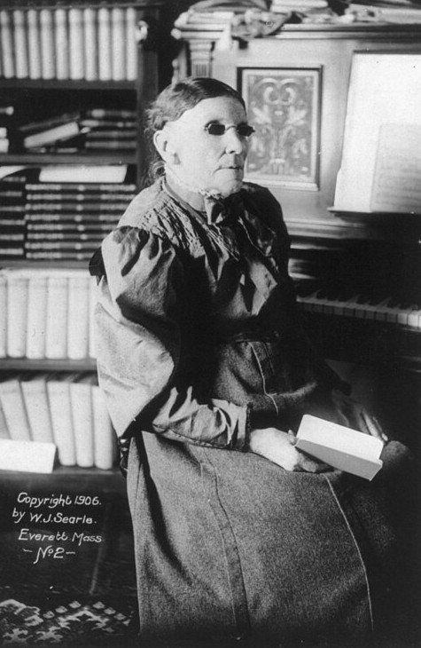 A black and white photo of Fanny J. Crosby, dated 1906, by W. J. Searle. Fanny is a woman in her eighties, wearing a long skirt and a blouse with puffed sleeves, and dark glasses. She is sitting at a piano, and holding a small book or booklet. There are several bookshelves in the background.