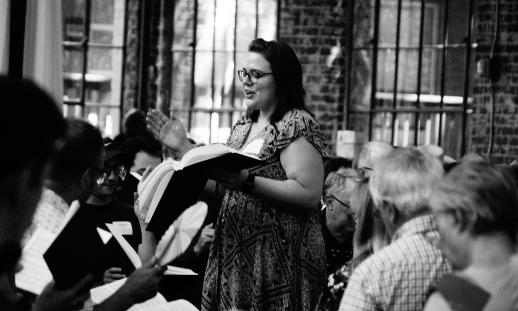 Vicki Elliott standing in the square, her right hand raised and her left hand holding The Sacred Harp 2025, as she leads her song 259 Easton at the United Convention in Atlanta, September 2025. Black and white photo by J Smith