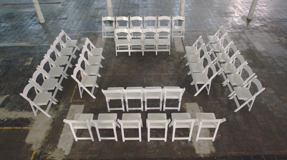 A screencap from Suzanne Lacy's film The Square and the Circle, showing an empty square of white folding chairs on the paved floor of the abandoned Brierfield Mill. There are white cast-iron columns supporting the next floor of the mill.