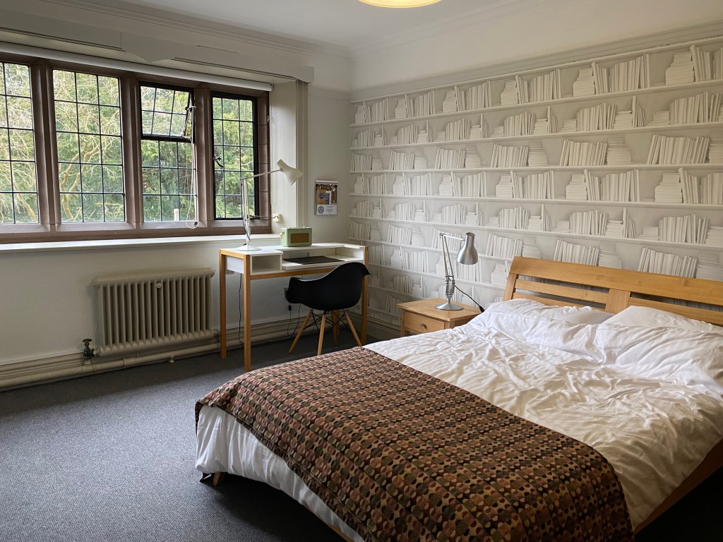 A double bed with rumpled covers and a low wooden headboard in a peaceful looking room. There is a window letting in light from the side, and a desk and chair positioned by the window.