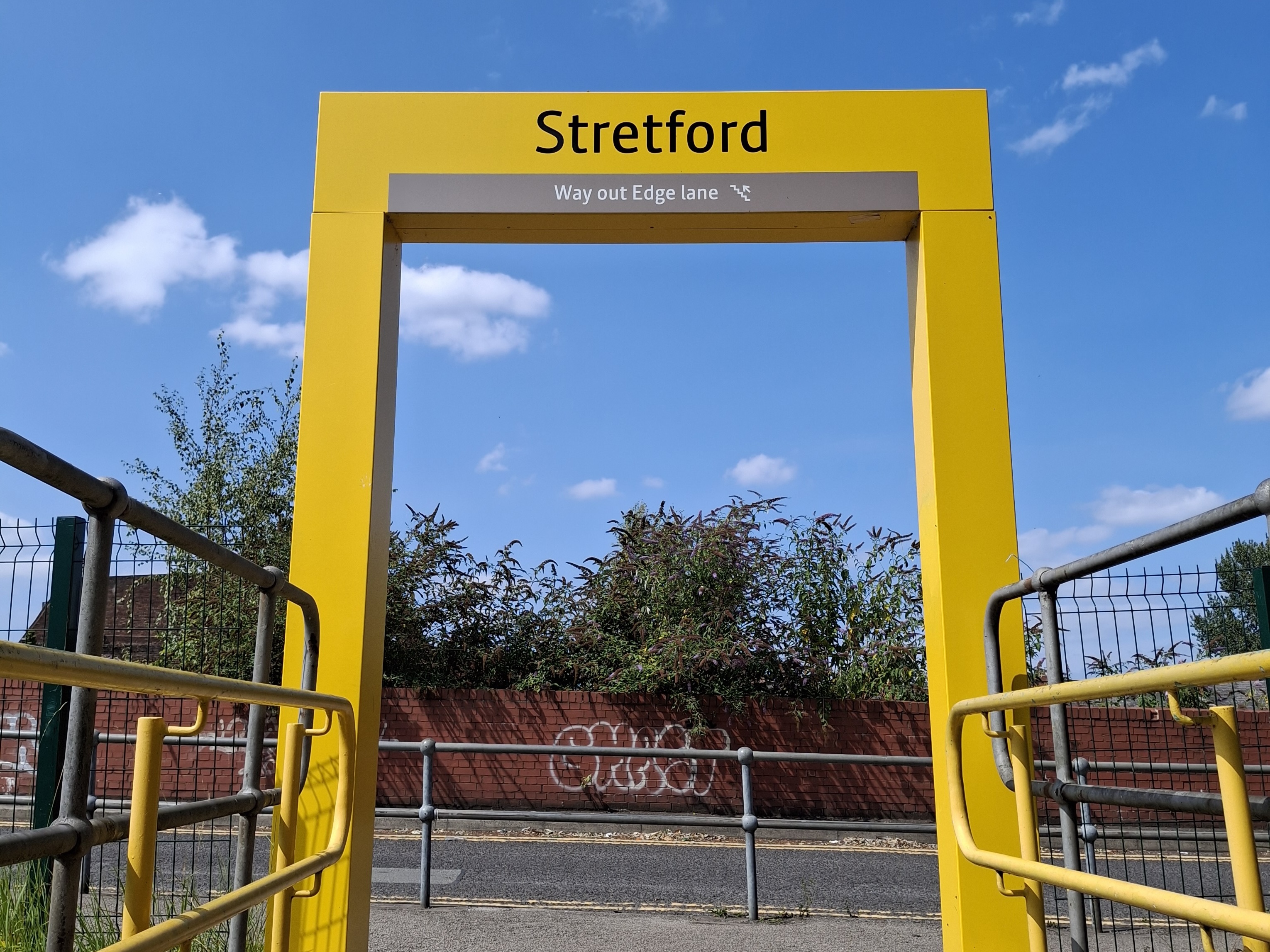 A vivid yellow arch towers above us against a bright blue summer sky. The place name 'Stretford' is in large black letters at the top. It's part of the Metrolink tram system's signage.