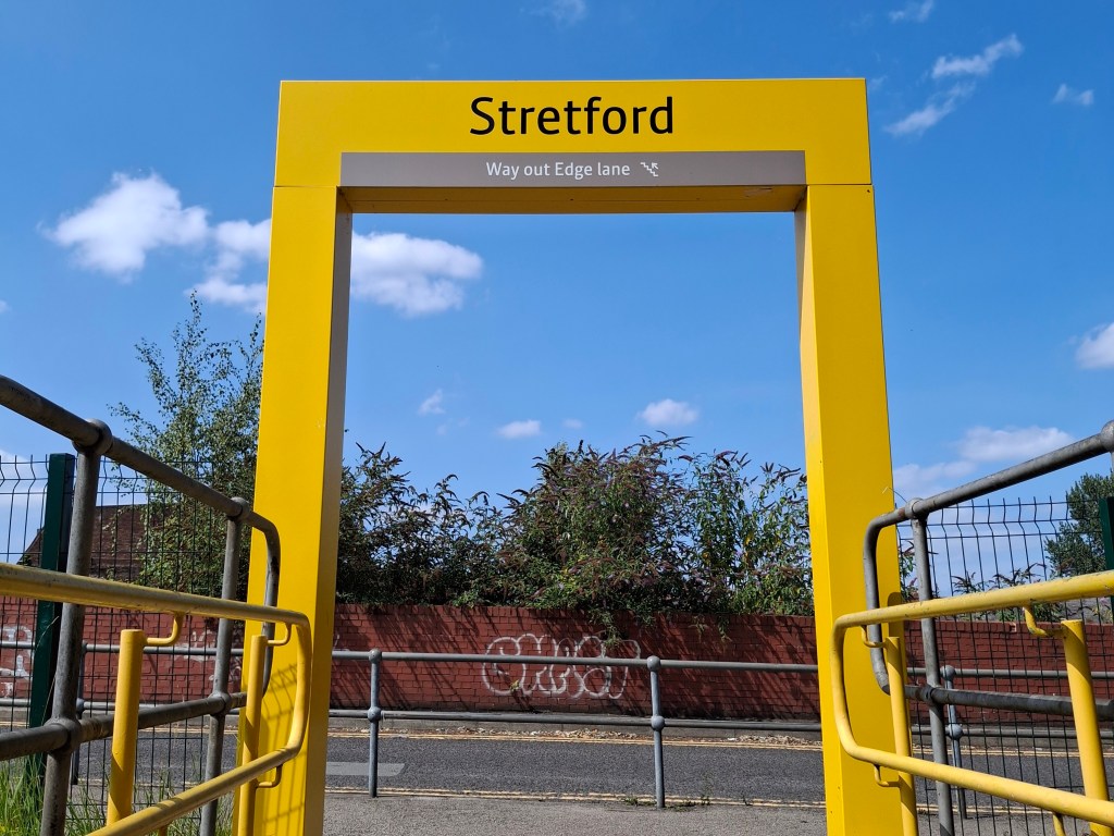 A vivid yellow arch towers above us against a bright blue summer sky. The place name 'Stretford' is in large black letters at the top. It's part of the Metrolink tram system's signage.