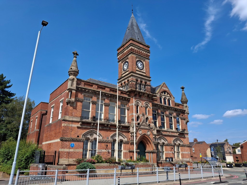 Stretford Public Hall - a large, impressive Victorian gothic building. It is mostly red brick with a grey slate roof and central clock tower over the grand arched entrance with a steeply pointed roof