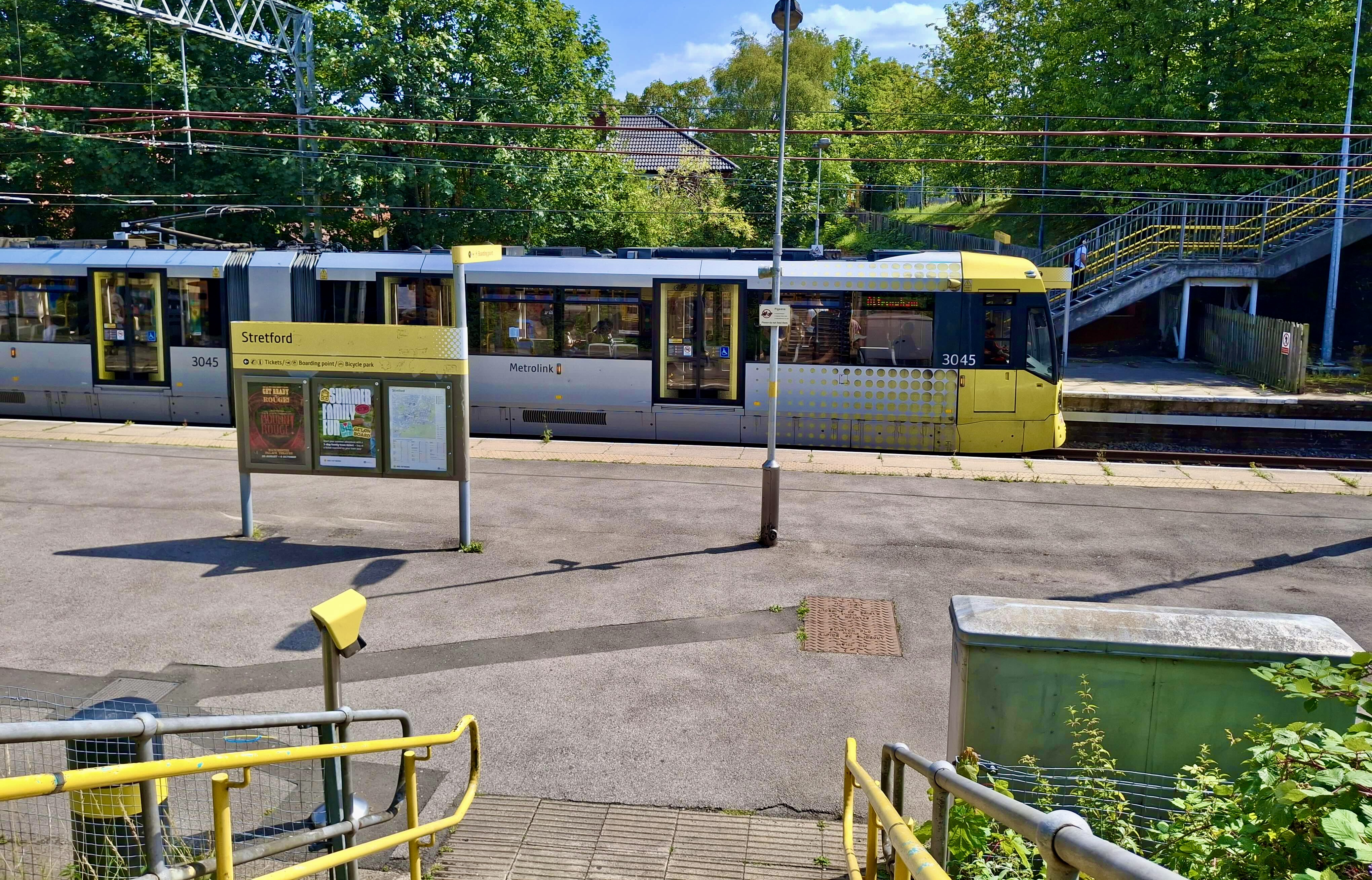 An image of a Metrolink tram taken just before going down the steps to the platform. It shows the side of the grey and yellow tram and the display board says Stretford.
