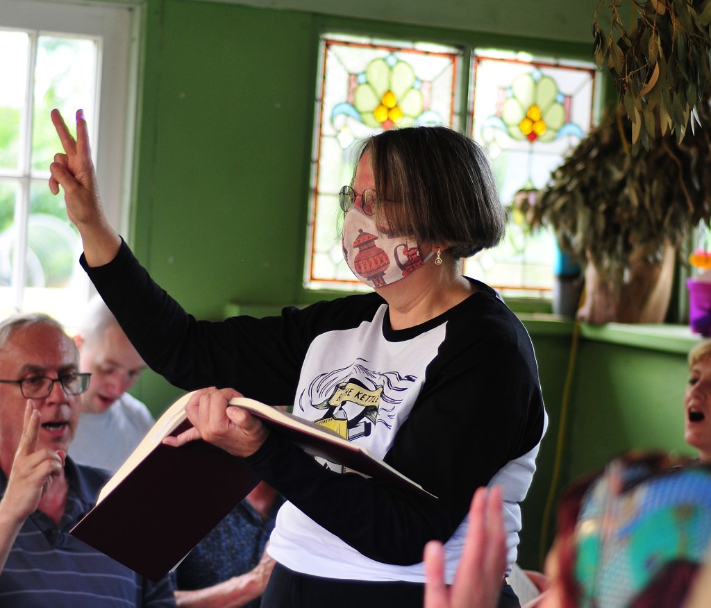 Photo by Susanna Cox: Harriet leading 178 Africa at the Frivolous Fields Folk Festival in June 2025 - standing in front of a green wall with a stained-glass window - the Frog House at Platt Fields, and wearing a mask, while signalling verse 2 with the right hand. Other singers are partly visible, some of them also signalling verse 2.