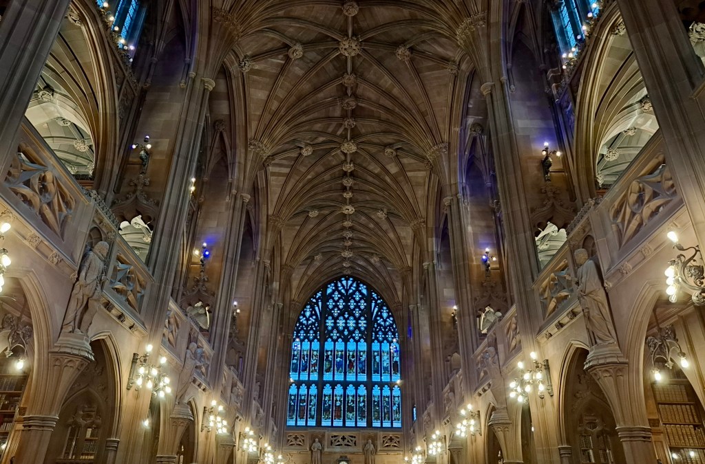 The spectacular ceiling of the Reading Room at John Rylands Library. It is vaulted and looks like a gothic cathedral. There is a large stained glass window at the end and statues around the sides of the upper floors. They look like saints, but are famous figures from literature, philosophy and art. 