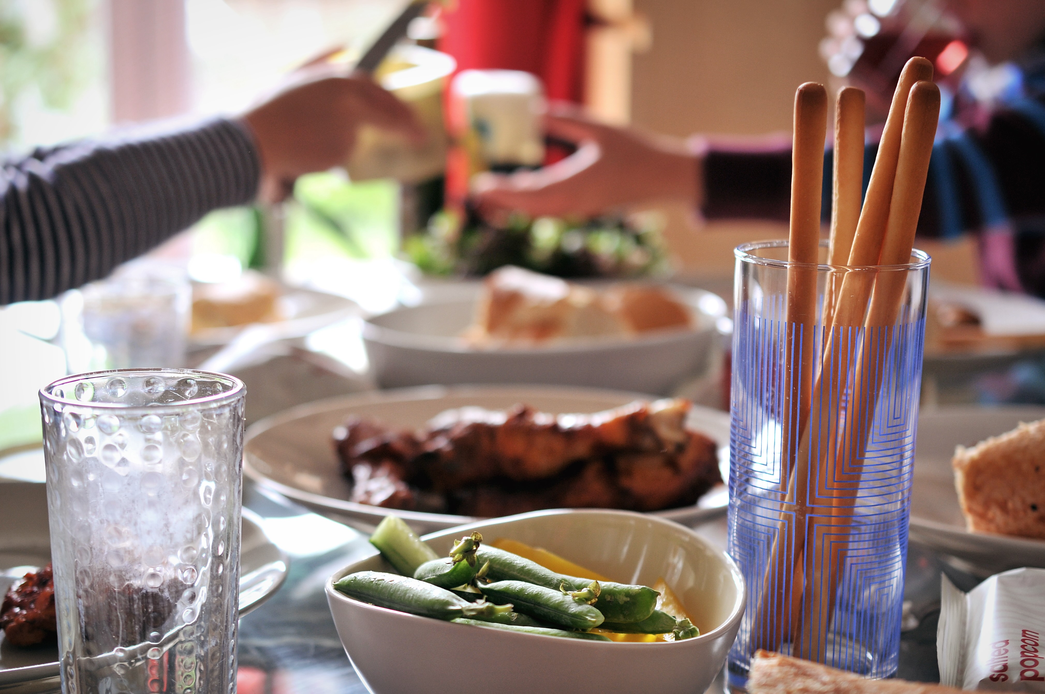Image of a shared meal. A table is generously laid with dishes of all kinds. There are bright green beans and breadsticks in a tall glass in the foreground. In the background, out of focus,  we see people's arms stretched across the table to exchange dishes. 