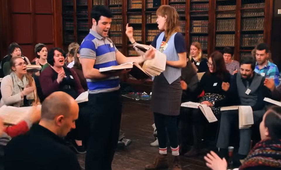 A screencap from the video of 47b Idumea, showing Irial Ó Cheallaigh (blue and grey striped shirt) and Claire Hogan (pale blue waistcoat) co-leading, with mostly altos and trebles visible behind them, and shelves of ancient leather-bound books in the background.