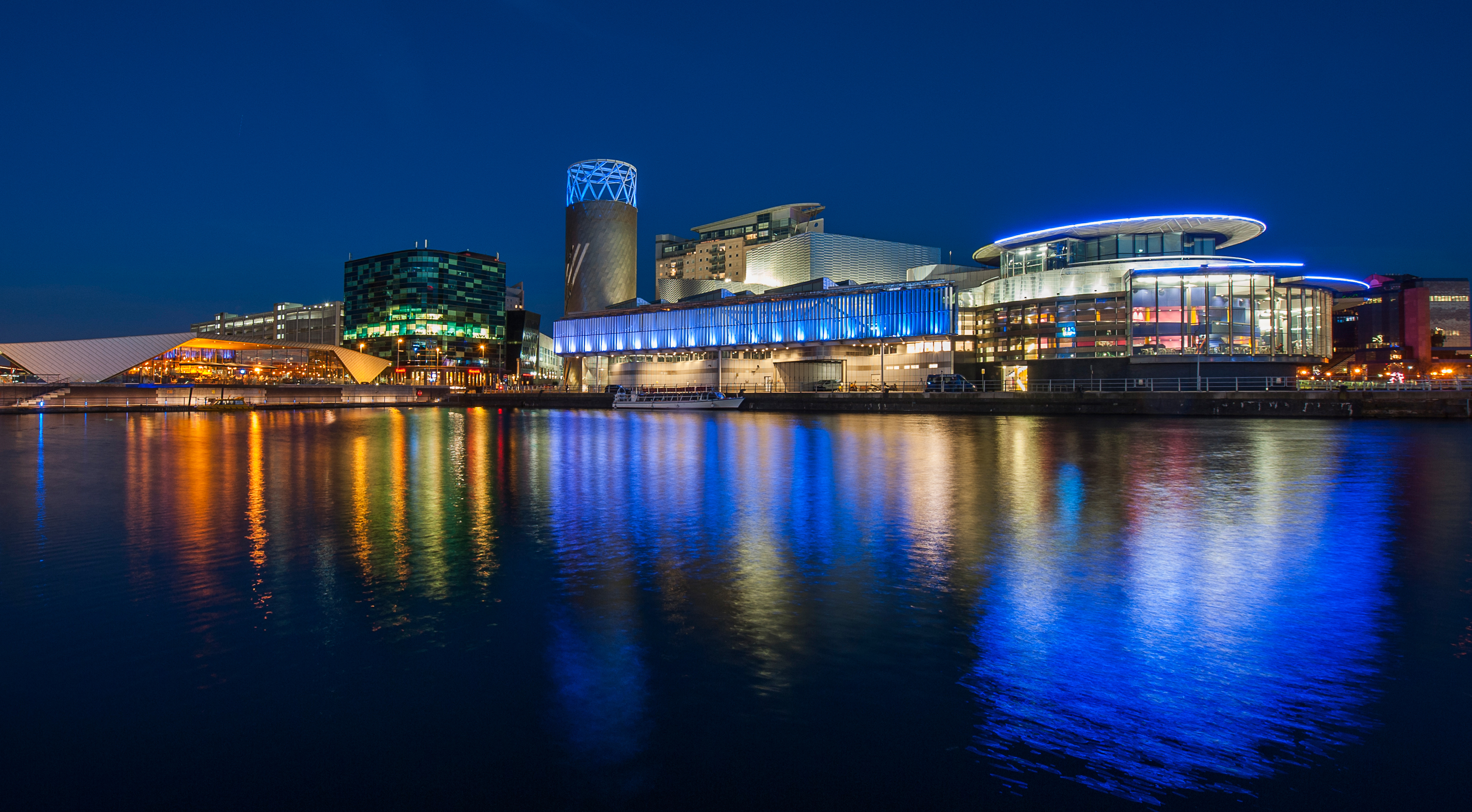 A night view across the water, with reflections of all the shiny metal buildings which are lit up by night. Each is a different colour - one electric blue, another green, another amber. 