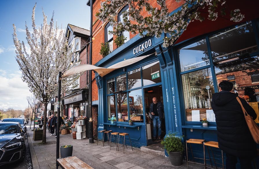 A photo of 'Cuckoo' bar in Prestwich on a spring day. The sky is pale blue, the building is striking red brick with large shiny windows and rich blue wooden window frames. Just outside, the street trees are covered in pale pink blossom. 
