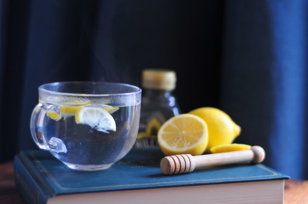 A clear glass cup with sliced lemon in steaming hot water stands on top of the dark green Sacred Harp book, along with a couple of bright yellow lemons and a carved wooden wand for drizzling honey. 