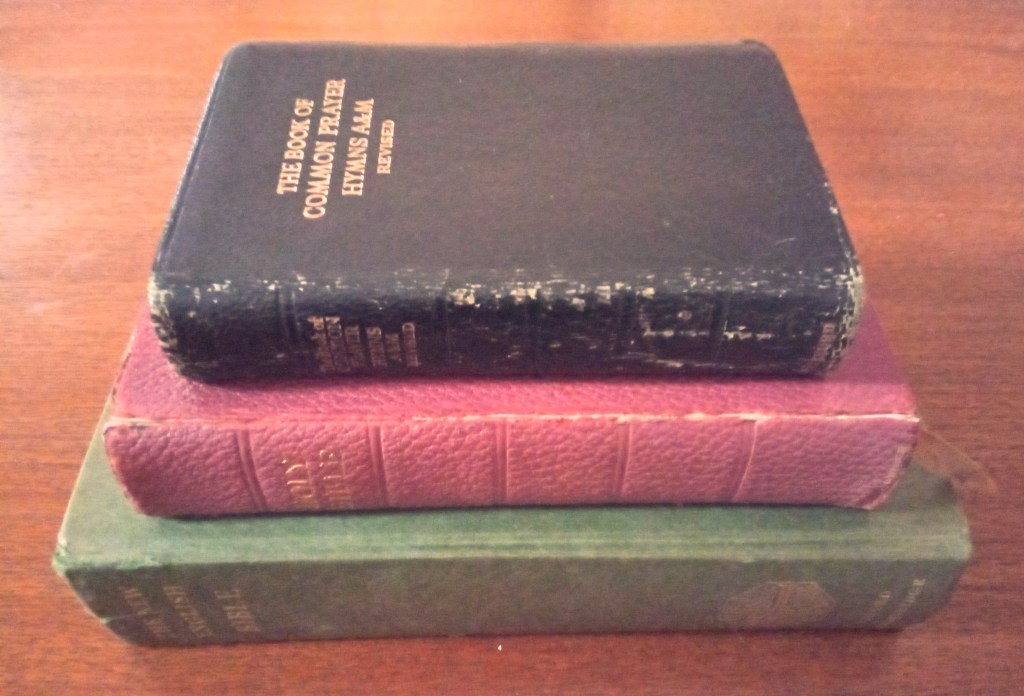 A pile of three rather battered books on a wooden table: at the bottom, a large green edition of the New English Bible, in the middle a reddish bound edition of the Authorised Version, labelled "Holy Bible", and on top a small black book combining the Book of Common Prayer and Hymns A&M Revised.