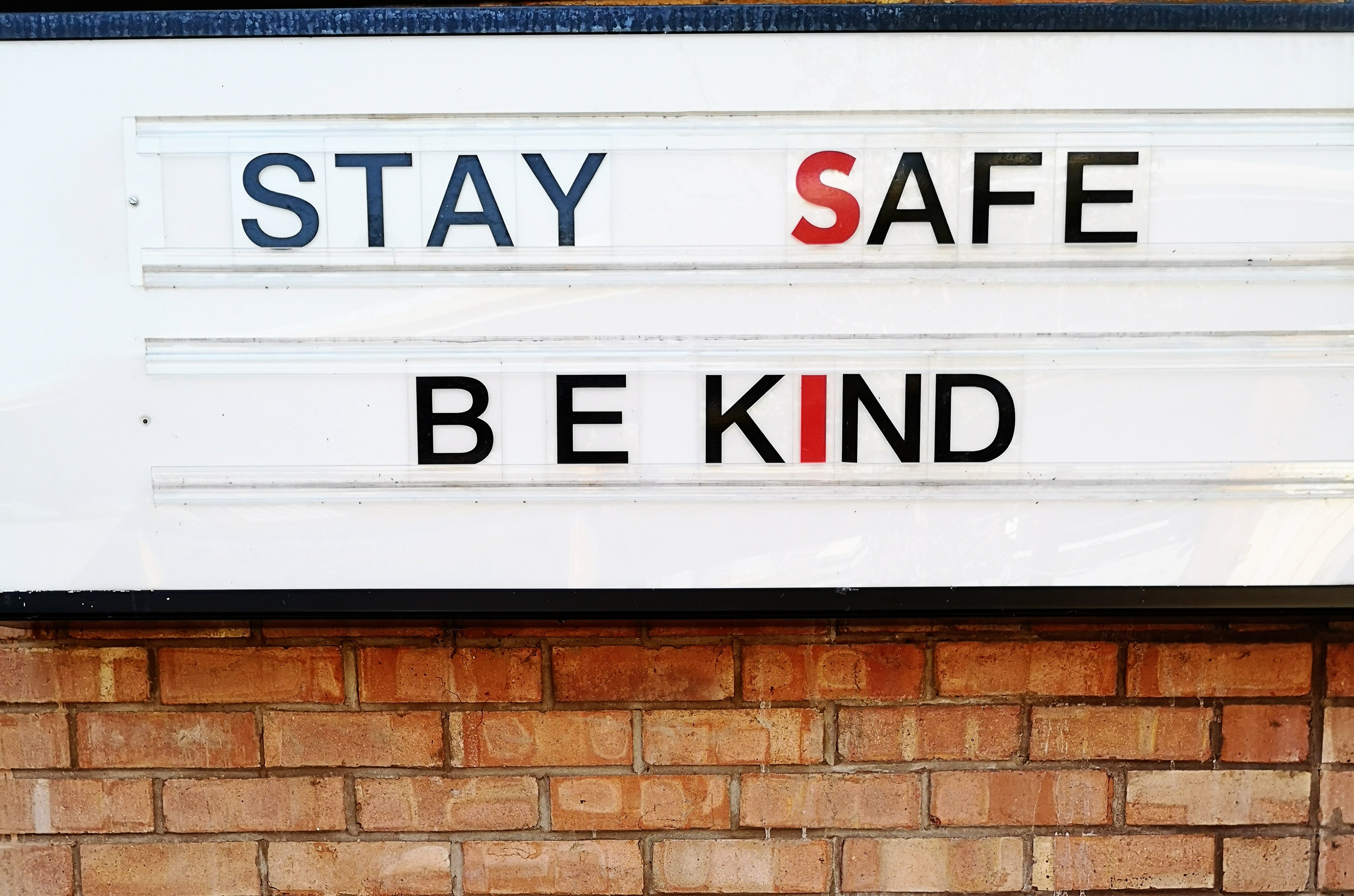 Photo of a sign on a red brick wall that reads STAY SAFE BE KIND in large black and red letter on a white background.