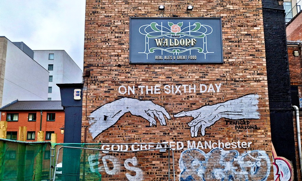 Image of a heavily graffitied end wall of a brown brick pub. A formal sign is printed 'The Waldorf - Real Ales & Great Food'. Underneath a street artist has painted outstretched male arms, with index fingers just touching (copied from Michelangelo's famous painting, 'Creation of Adam'). The caption reads 'On the Sixth Day God Created MANchester'