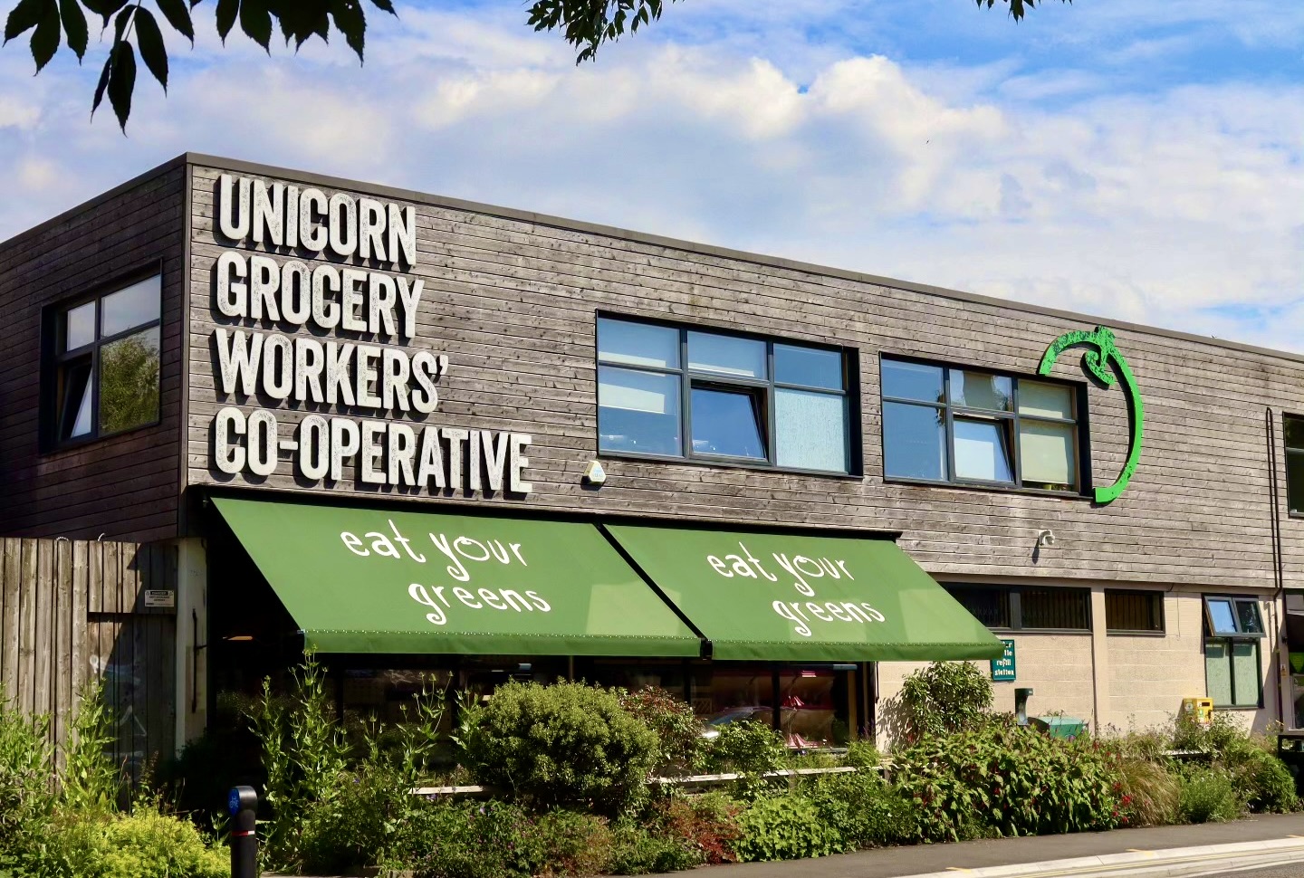 A long low building that is cladded with brown wooden planks. In large white letters on the building it says Unicorn Grocery Workers' Co-operative.  Two green canopies have the slogan 'Eat your greens' on them. In front there are shrubs and grasses growing. 