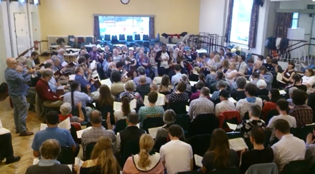 A screencap from the 2014 UK convention at Stannington in Yorkshire. There are about 150 singers in the square, with Jenneith Codrington standing in the middle to lead.
