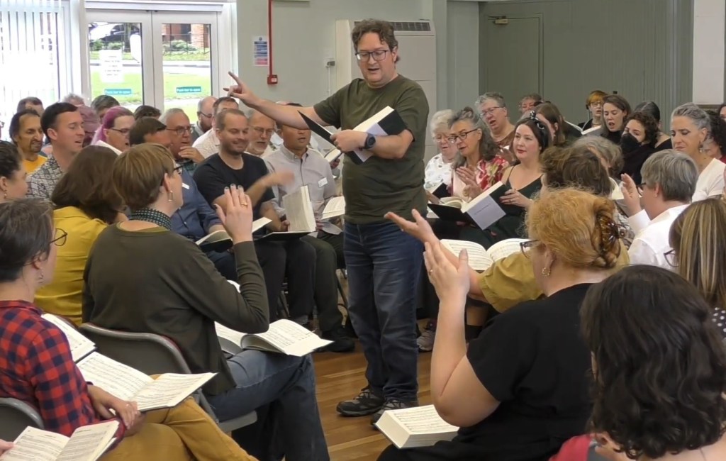 Screencap of Jan leading 524 Moonlight at the UK convention in September 2025. He is clean-shaven and wearing a green Tshirt, perhaps to match the new Sacred Harp edition. His right arm is raised, and singers all around the square are looking up to follow his lead. Video by Werner Ullah and James Harvey.