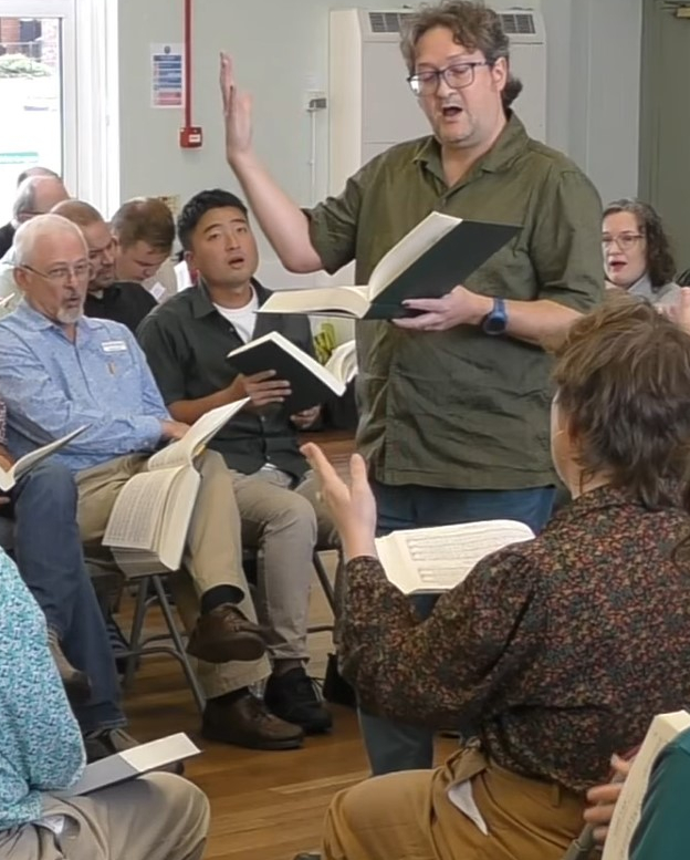 Screencap of Jan leading 70t Gainsville at the UK convention in Barley Mow near Durham in September 2025. He is clean-shaven and wearing a green shirt, perhaps to match the new Sacred Harp edition. His right arm is raised, and he holds the book in his left hand. The screencap is from the video by Werner Ullah and James Harvey.